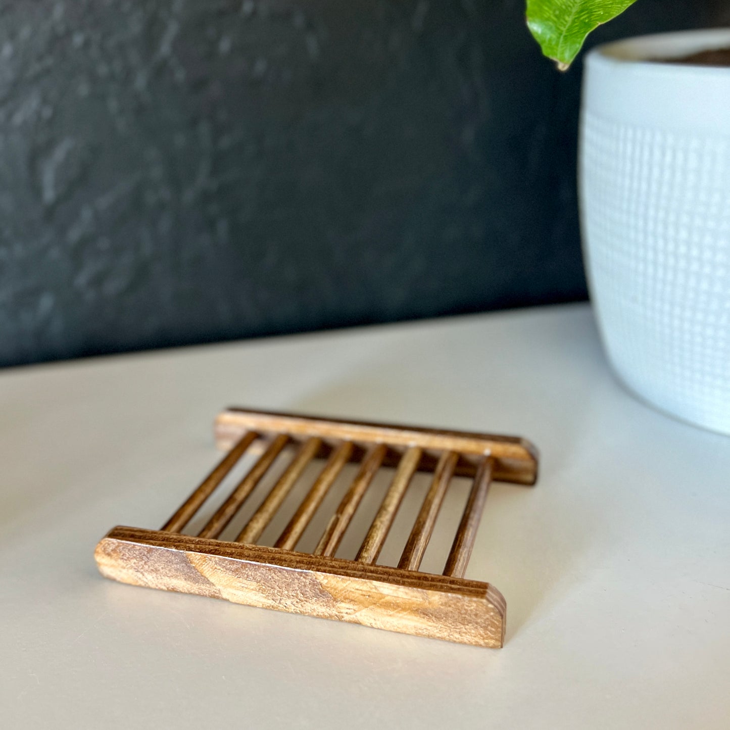 A bamboo soap dish is being displayed. It is a charcoal bamboo color. It is on a shelf with a black wall background and a white plant pot with a plant leaf in the corner.