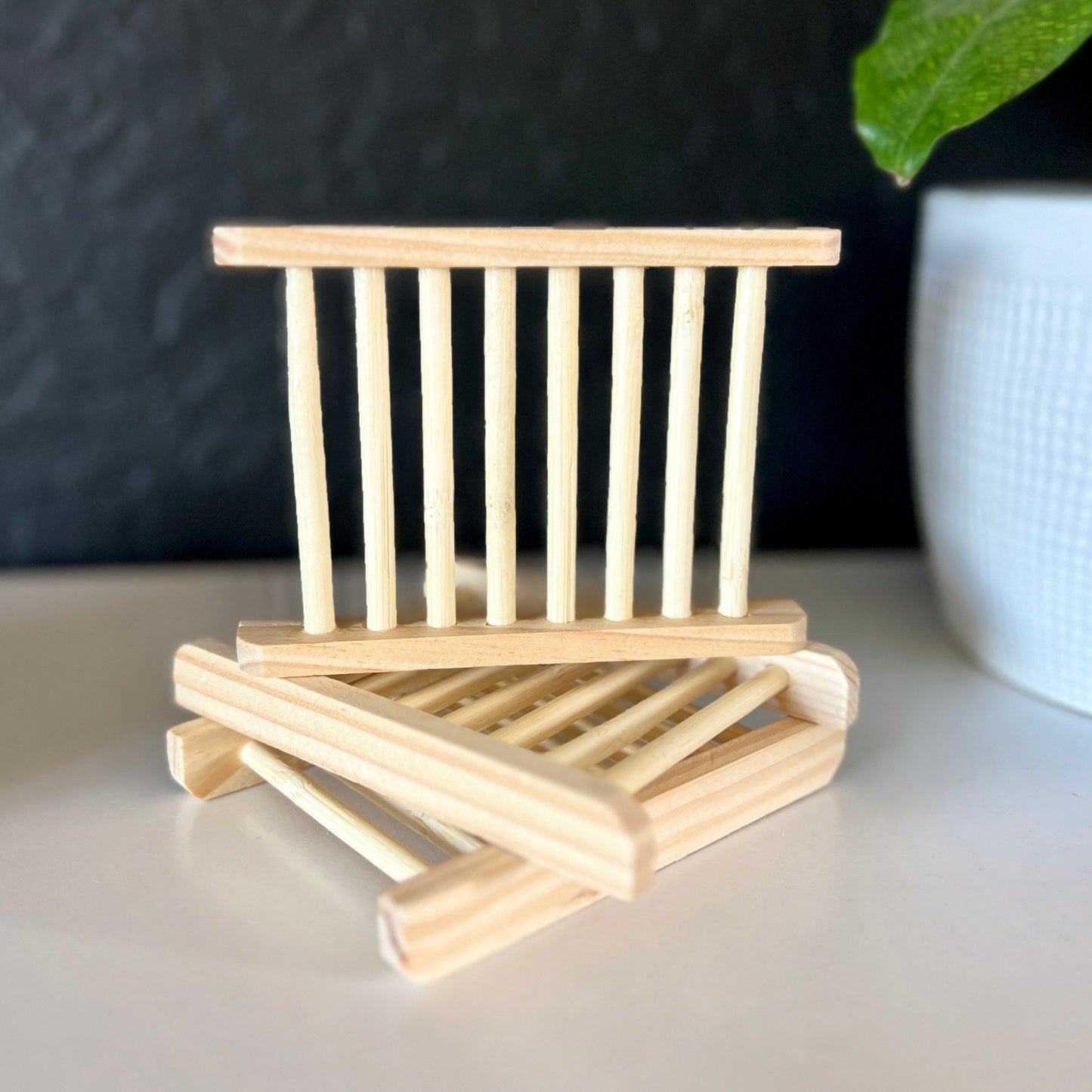 A bamboo soap dish is being displayed on two other soap dishes. It is a natural bamboo color. It is on a shelf with a black wall background and a white plant pot with a plant leaf in the corner.