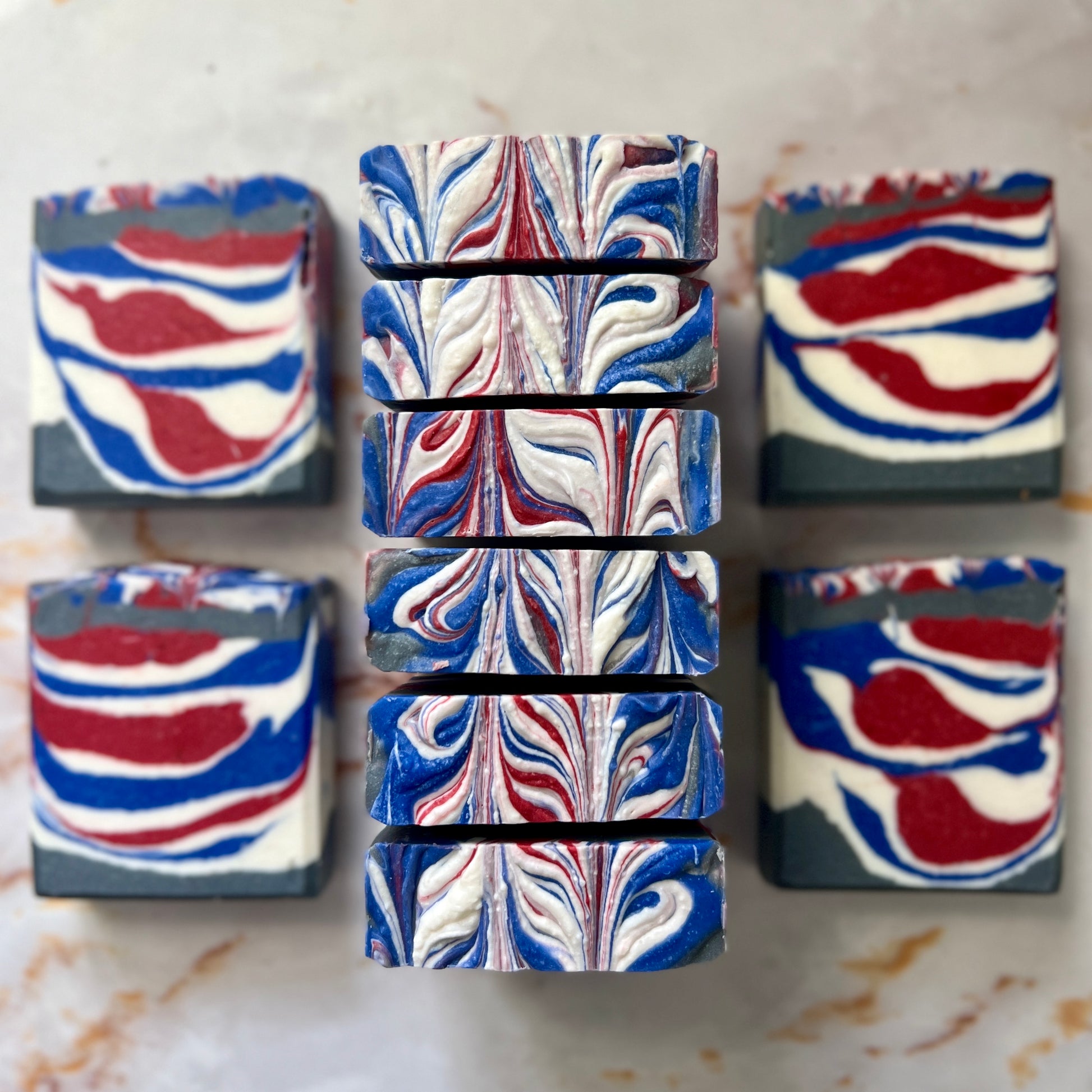 Handmade soap on a white countertop showing the top and front of the soap. It is a grey, white, blue, and red soap. The colors are layered and alternating on top of each other to create a line design. 