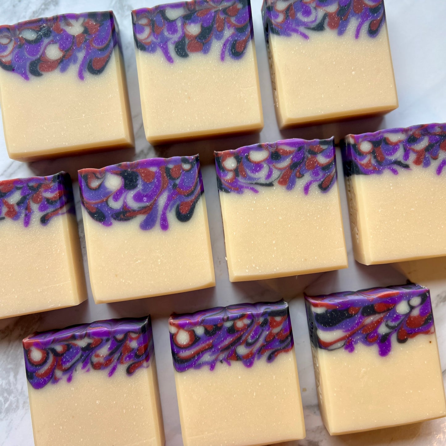 Handmade soap on a countertop lined up to show the design. Cream colored base with various shades of purples and red that were poured into the base color.