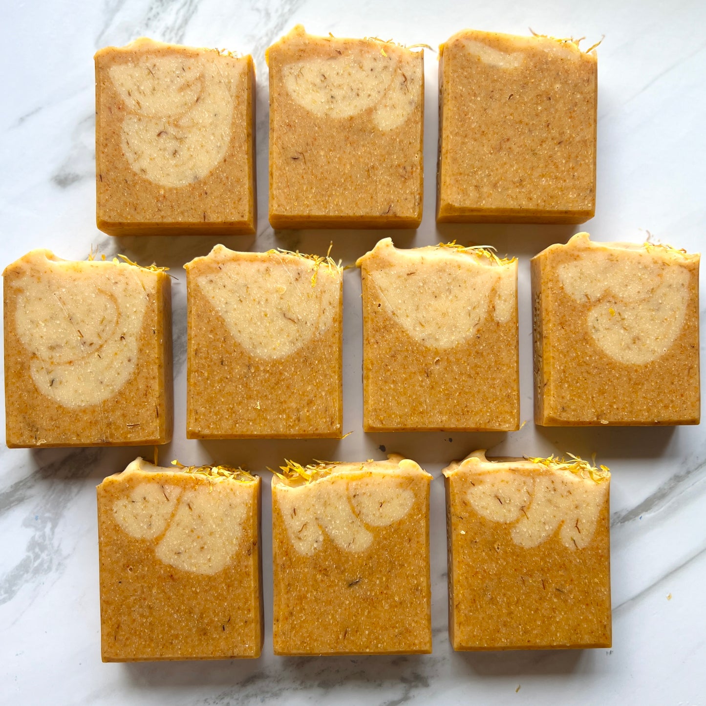 Handmade soap on a countertop showing the front of the soap. Orange soap with speckles with a lighter orange with speckles dropped into the soap creating swirls. The top is textured with large lines and calendula petals on top.