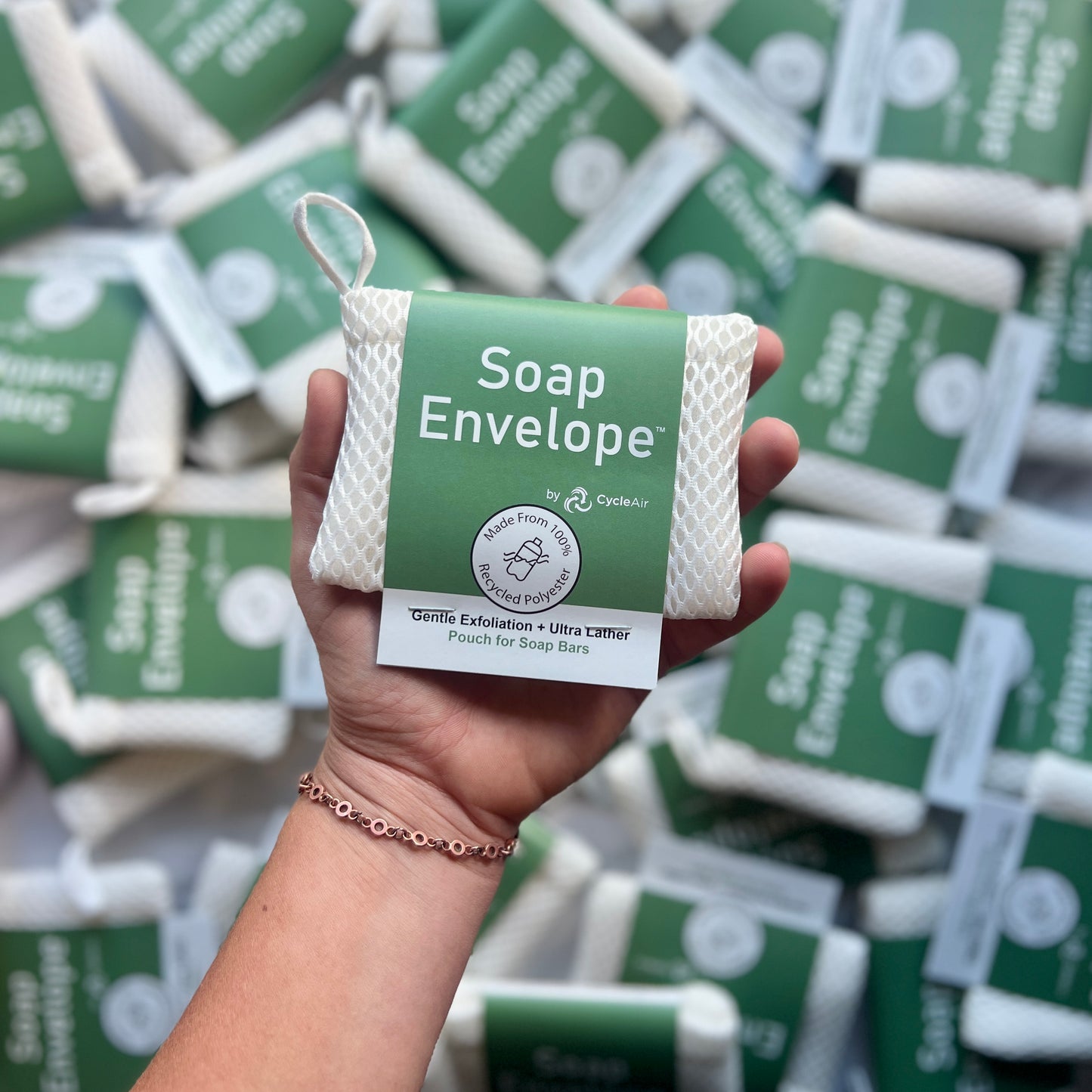 White soap envelope with green packaging being held in a hand wearing a bracelet above other soap envelopes on a desk.