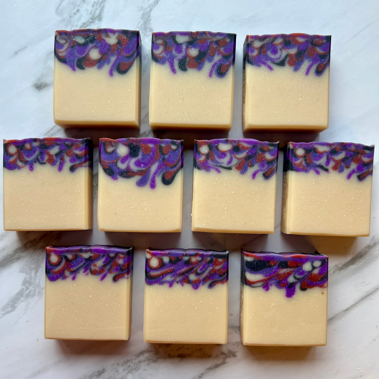 Handmade soap on a countertop lined up to show the design. Cream colored base with various shades of purples and red that were poured into the base color.