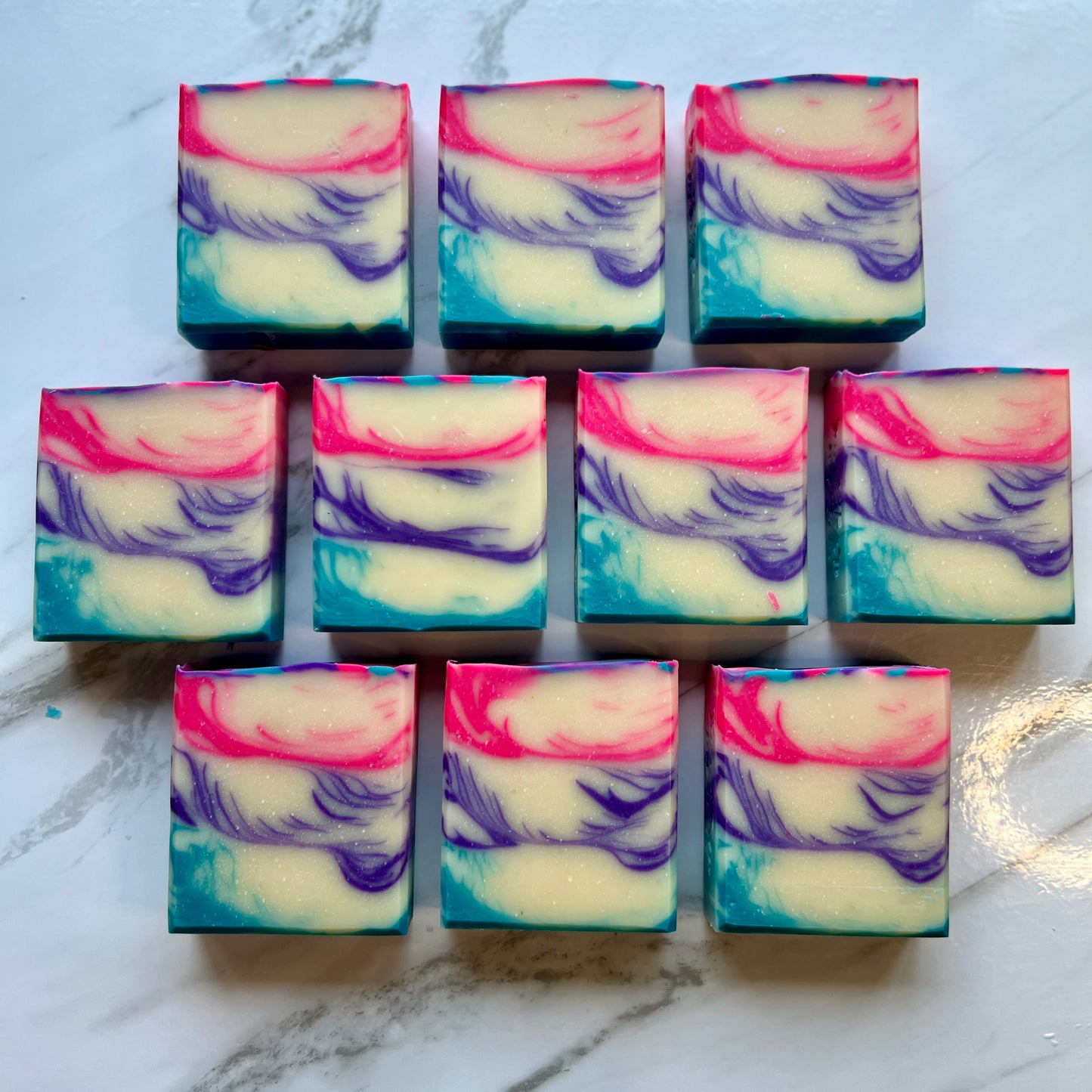 Handmade soap on a countertop showing the top and front of the soap. White colored base with a blue accent on the bottom poured with lines forming. The middle accent color is purple with lines forming. The top layer is the same design with a pink color.