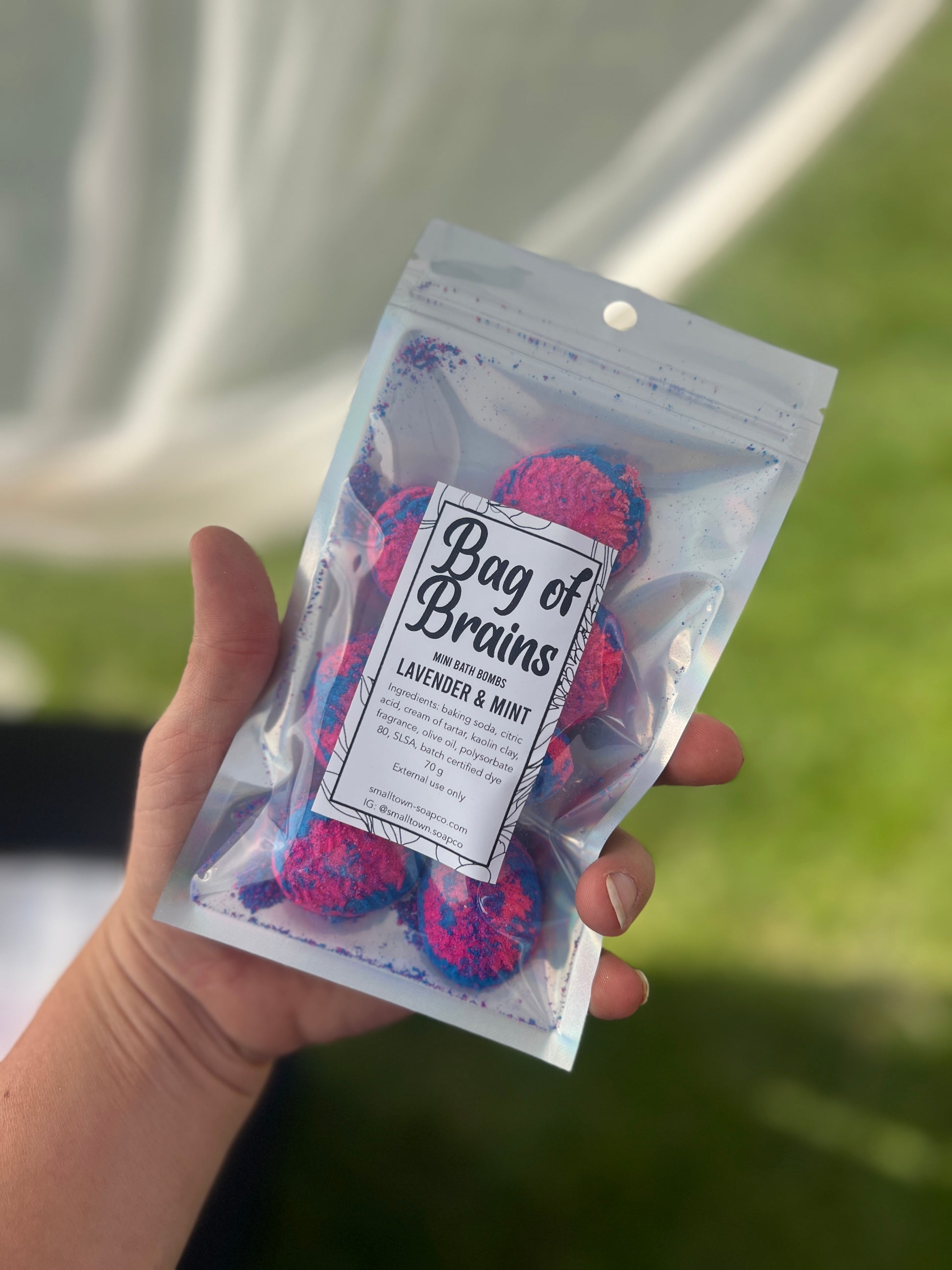 A holographic bag with blue and pink mini brains bath bombs with a label on top of the bag. The bag has a see through front. The bag is being held by a hand with a with curtain and grass in the background.