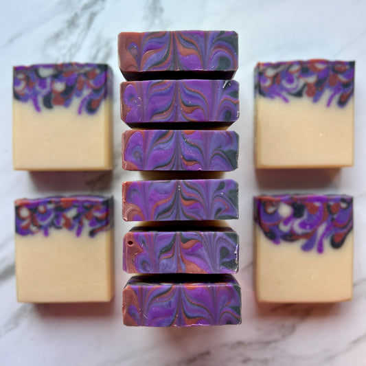 Handmade soap on a countertop showing the top and front of the soap. Cream colored base with various shades of purples and red that were poured into the base color.