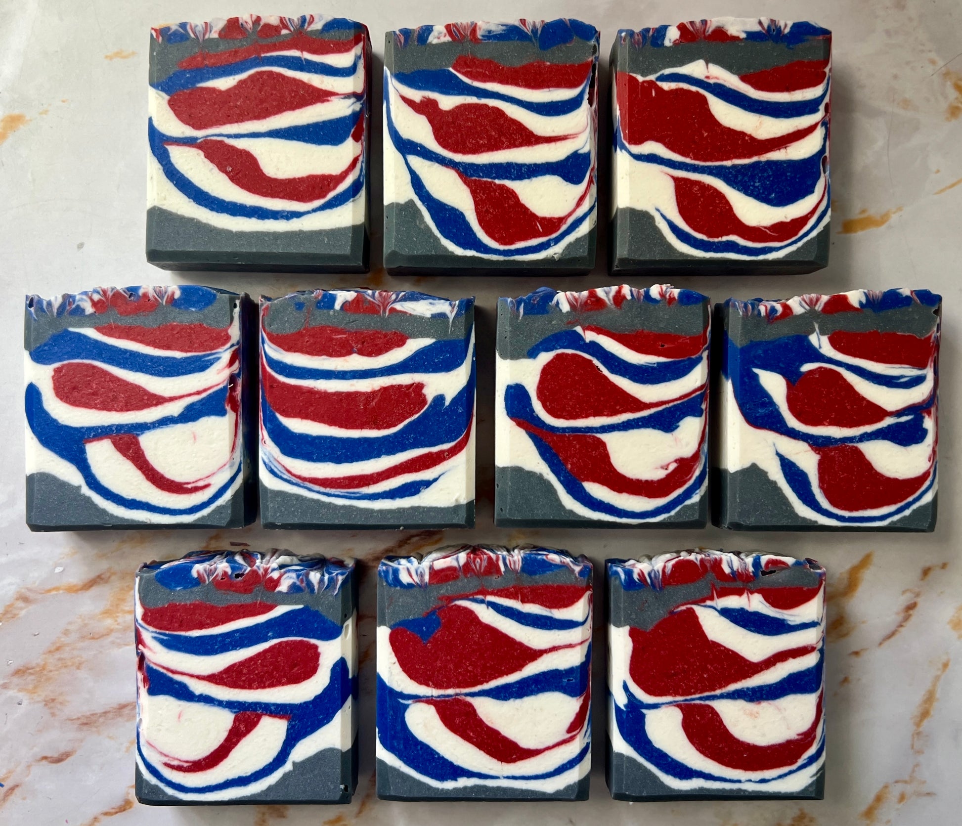 Handmade soap on a white countertop showing the front of the soap. It is a grey, white, blue, and red soap. The colors are layered and alternating on top of each other to create a line design. The soaps are lined up to show their design.