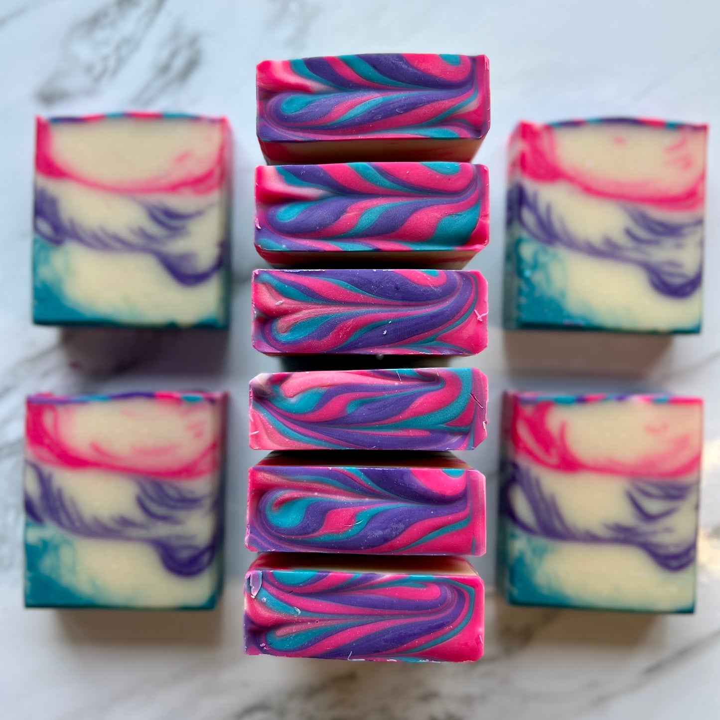 Handmade soap on a countertop showing the top and front of the soap. White colored base with a blue accent on the bottom poured with lines forming. The middle accent color is purple with lines forming. The top layer is the same design with a pink color. The top is pink purple and blue textured.