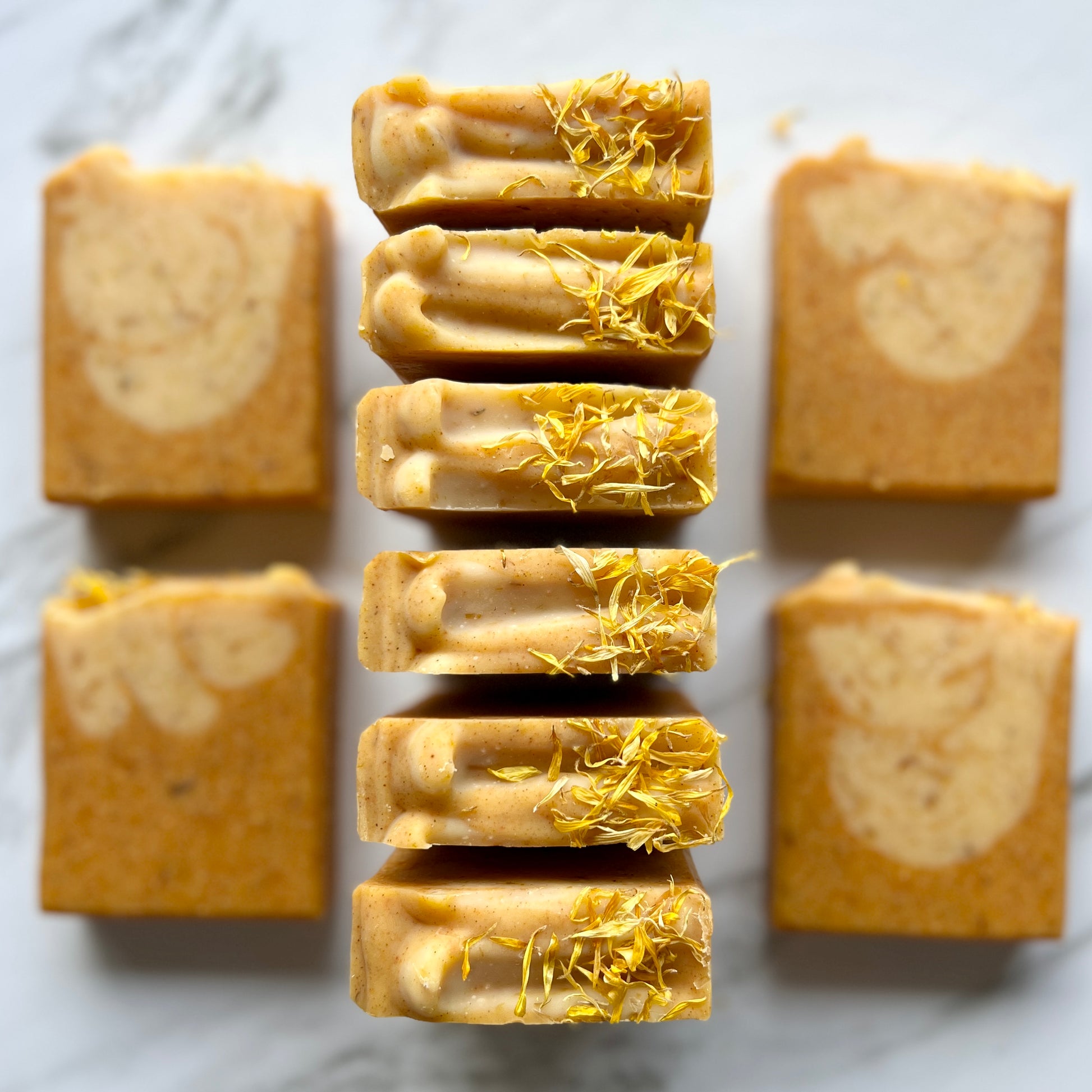 Handmade soap on a countertop showing the top and front of the soap. Orange soap with speckles with a lighter orange with speckles dropped into the soap creating swirls. The top is textured with large lines and calendula petals on top.