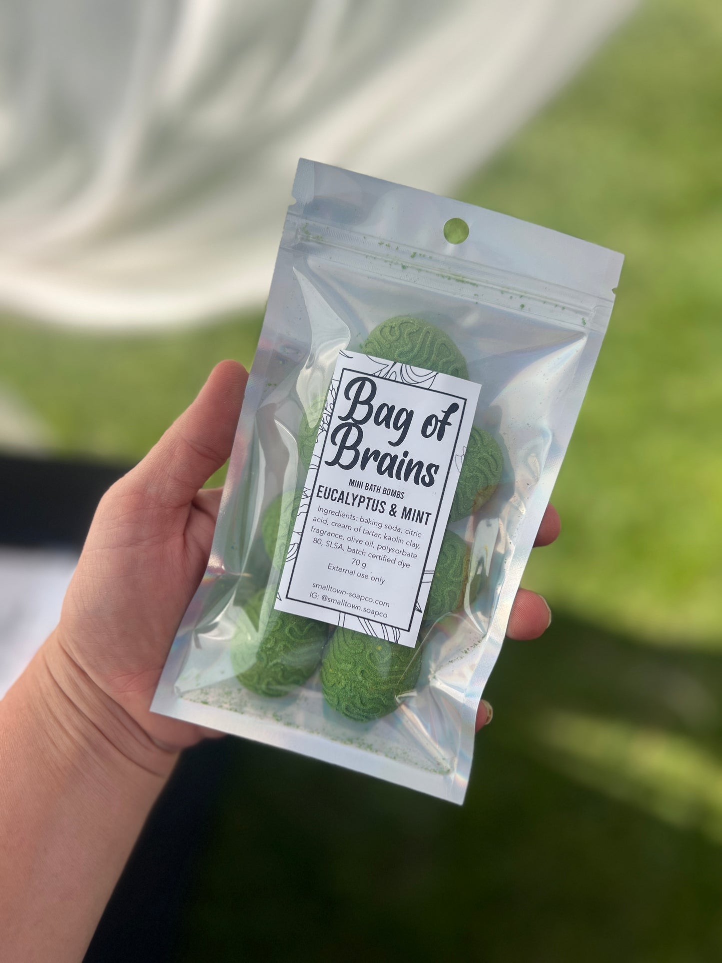 A holographic bag with green mini brains bath bombs with a label on top of the bag. The bag has a see through front. The bag is being held by a hand with a with curtain and grass in the background.