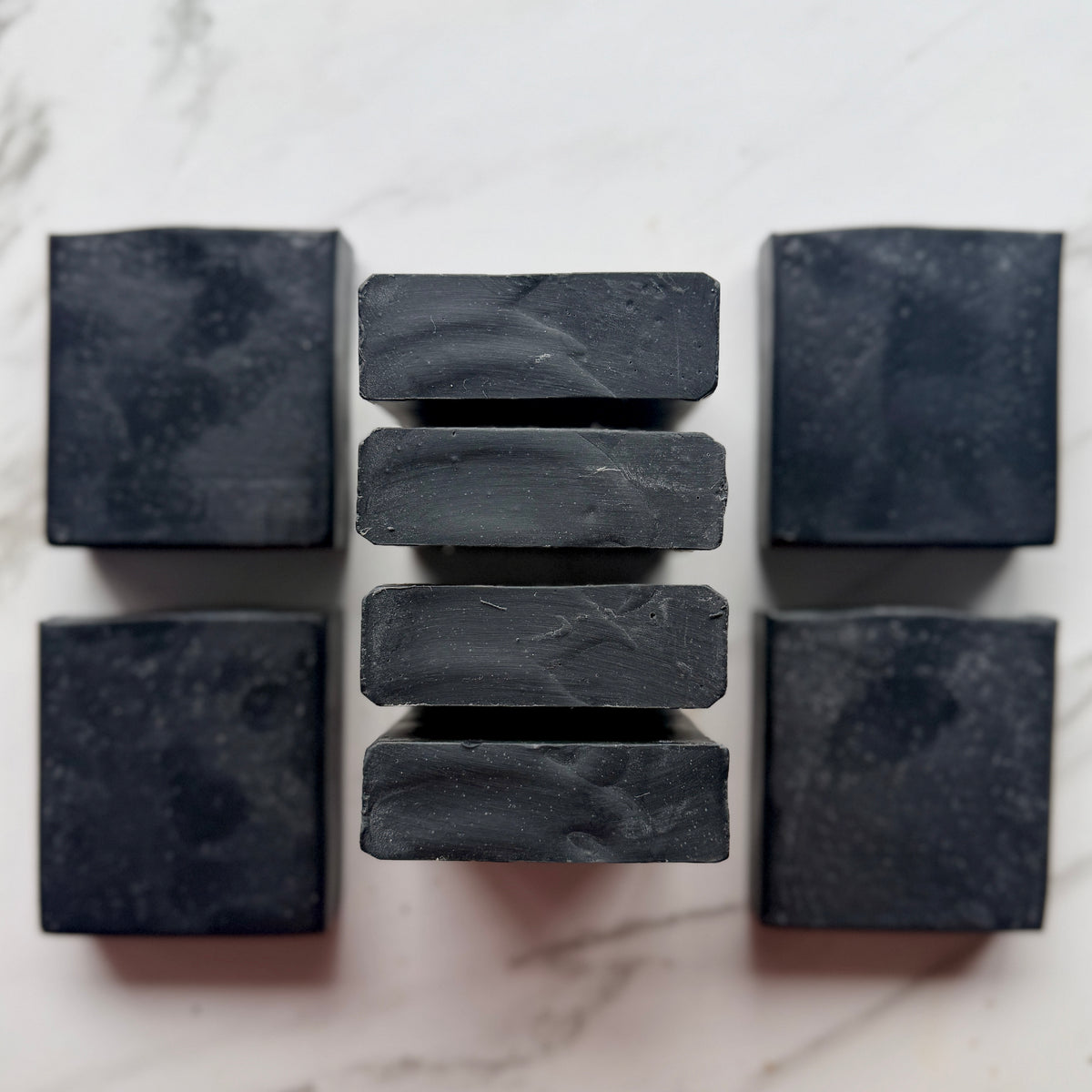 Eight dark charcoal gray rectangular soap bars arranged symmetrically on a light marble surface, with four stacked in the center and two standing on either side.