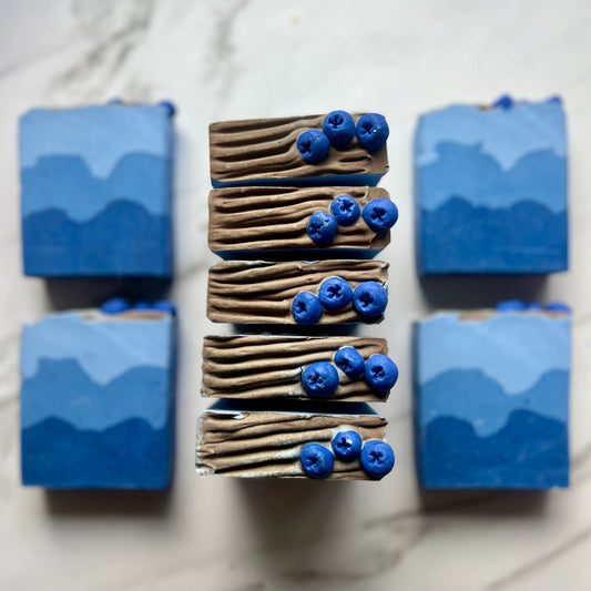 Handmade soap on a countertop showing the top and front of the soap. Blue soap with three layers. Dark blue on the bottom, a lighter shade in the middle, and the lightest blue on top. The top is discolored to a grey ish brown and is textured with lines. There are soap dough embers on the top to look like blueberries. 3 soap dough blueberries per soap. 