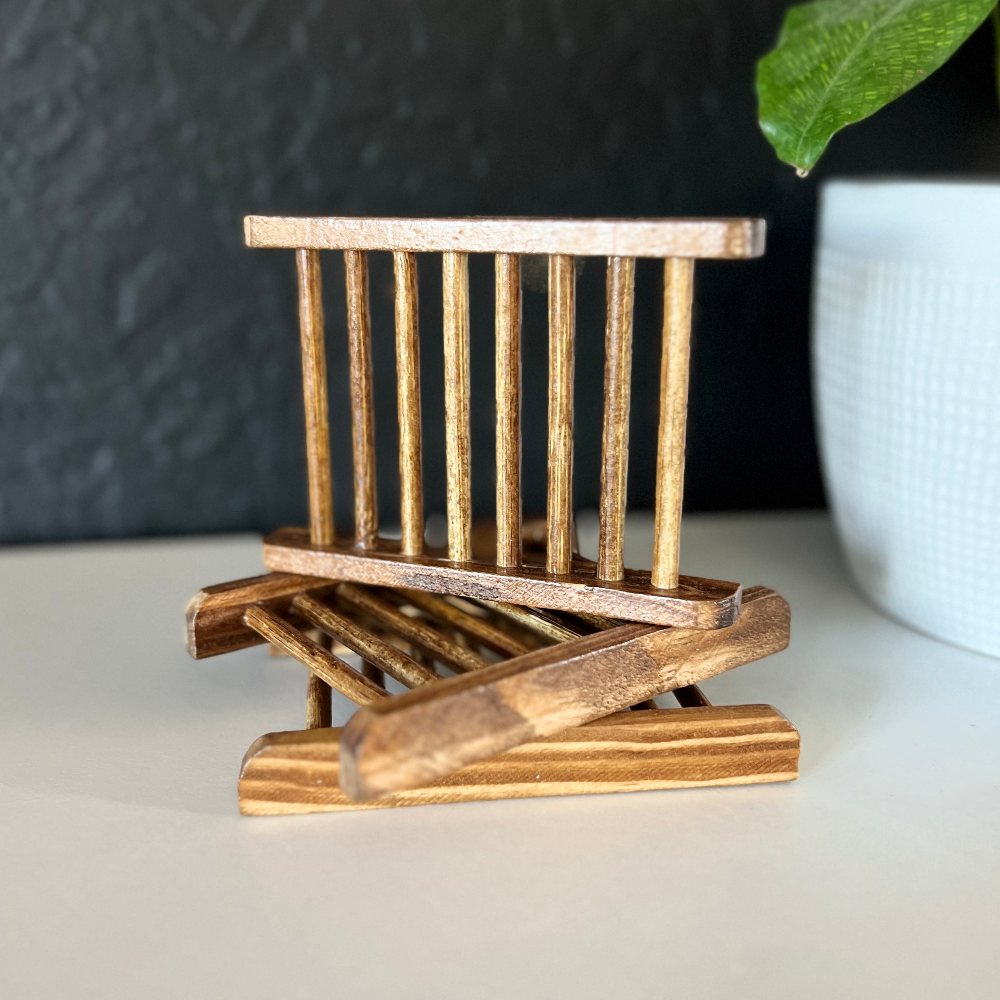 A bamboo soap dish is being displayed on two other soap dishes. It is a charcoal bamboo color. It is on a shelf with a black wall background and a white plant pot with a plant leaf in the corner. 