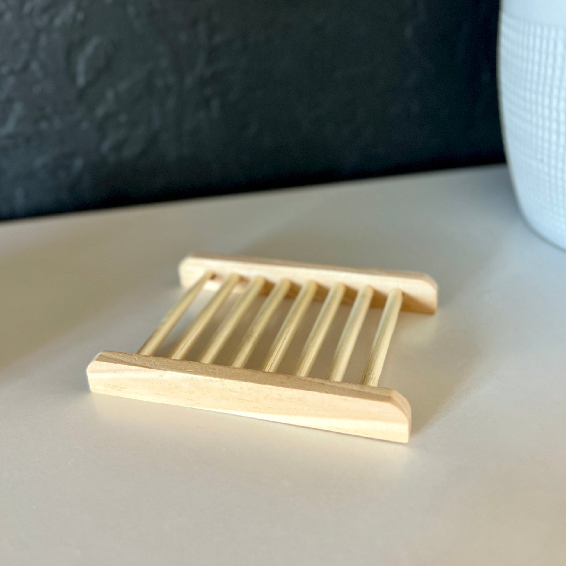 A bamboo soap dish is being displayed. It is a natural bamboo color. It is on a shelf with a black wall background and a white plant pot in the corner.